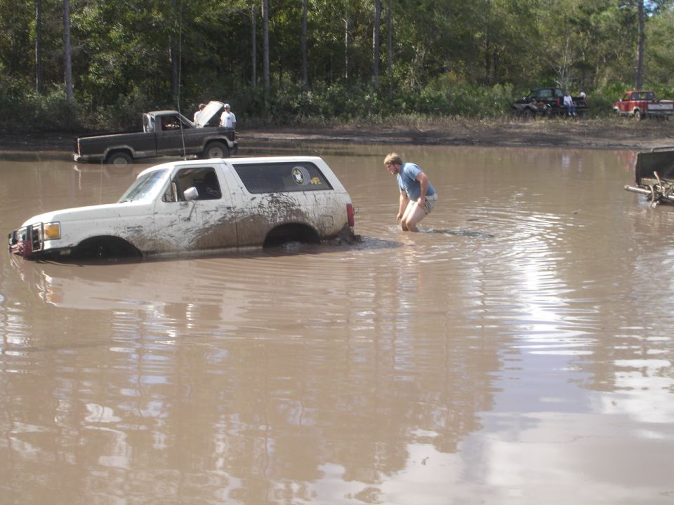 White SUV stuck in muddy water, side view. Mud splattered on body. A person walks nearby