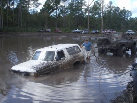 1989 Ford Bronco - A white SUV is submerged in a muddy water hole while a person attempts to assist.