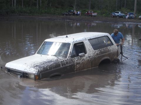 1989 Ford Bronco - A white SUV is stuck in a mud pit, with a person trying to extract it.