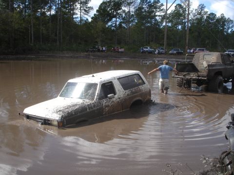1989 Ford Bronco - A white SUV is submerged in a mud pit while a man walks through the water towards another vehicle.