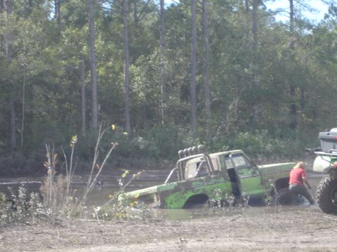 1989 Ford Bronco - A person attempts to free a green pickup truck that is stuck deep in the mud, with an ATV nearby.