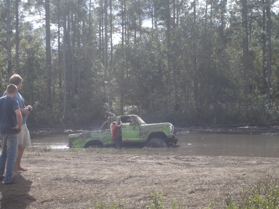 Green pickup truck stuck in mud, side view. Two people stand on the bank
