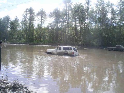 1989 Ford Bronco - White 1989 Ford Bronco is stuck in a muddy waterhole, with another truck visible in the distance.