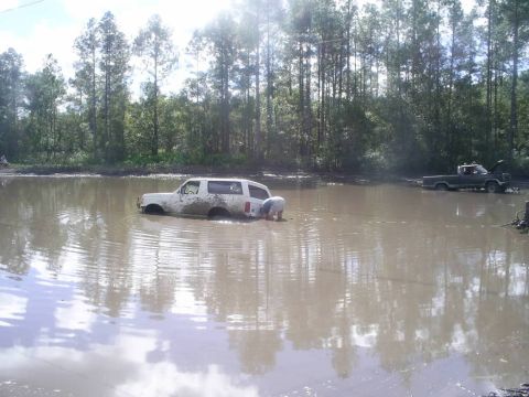 1989 Ford Bronco - A white 1989 Ford Bronco is submerged in muddy water, with one person working on it and another truck visible in the background.