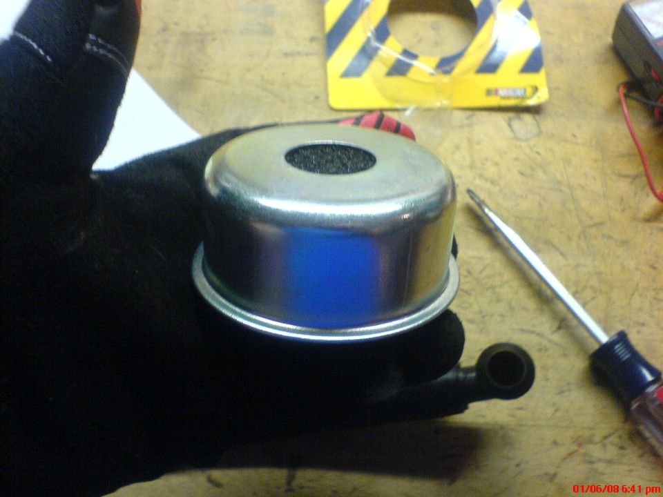 Gloved hand holding a chrome oil breather cap with foam filter over a workbench with screwdriver and packaging
