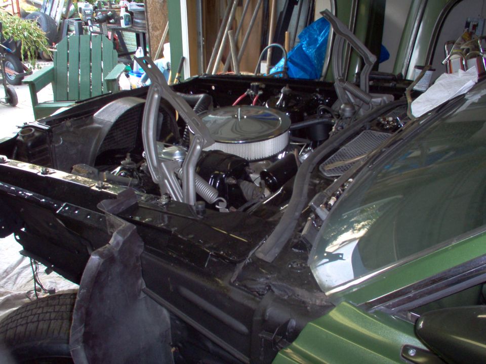 Car engine bay with hood open, showing engine, air filter, and parts of the chassis. Another car is partially visible on the right
