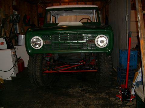 1974 Ford Bronco - A green 1974 Ford Bronco with a white roof is parked inside a garage, showing its front grille and lifted suspension.