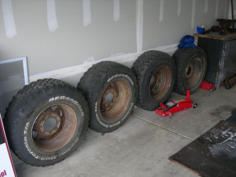 1985 Ford Bronco - Four BFGoodrich Mud-Terrain T/A tires on rusty wheels are lined up next to a red floor jack.