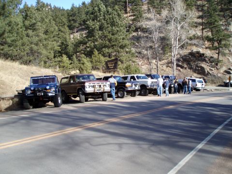 1979 Ford Bronco - Several SUVs and trucks, including a 1979 Ford Bronco, are parked along a road with a wooded hillside.