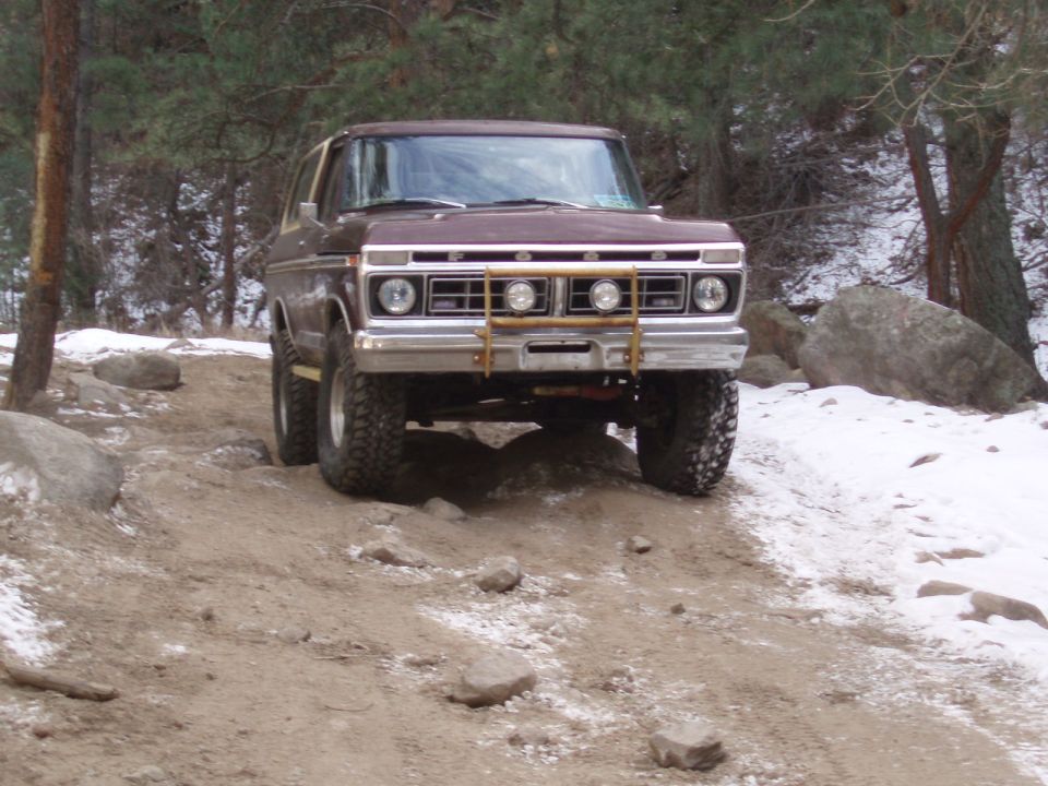 Brown 1978 Ford Bronco with a brush guard and off-road tires driving on a dirt path with snow