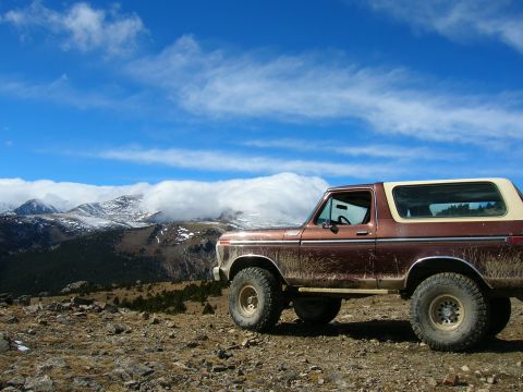 1979 Ford Bronco - A brown 1979 Ford Bronco sits on a rocky overlook with snow-capped mountains in the background under a blue, cloudy sky.