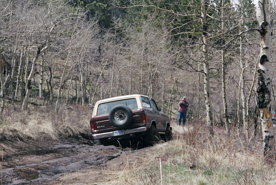 Maroon and white Ford Bronco with spare tire on rear, driving on muddy trail