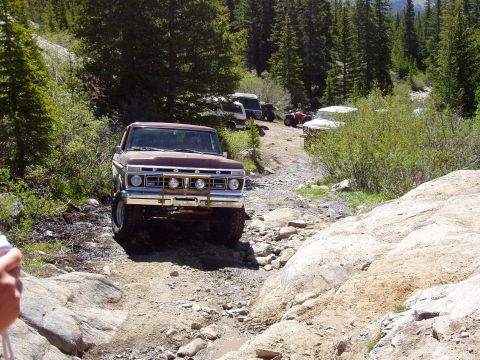 1979 Ford Bronco - A lifted Ford truck navigates a rough, off-road path with trees and other vehicles nearby.