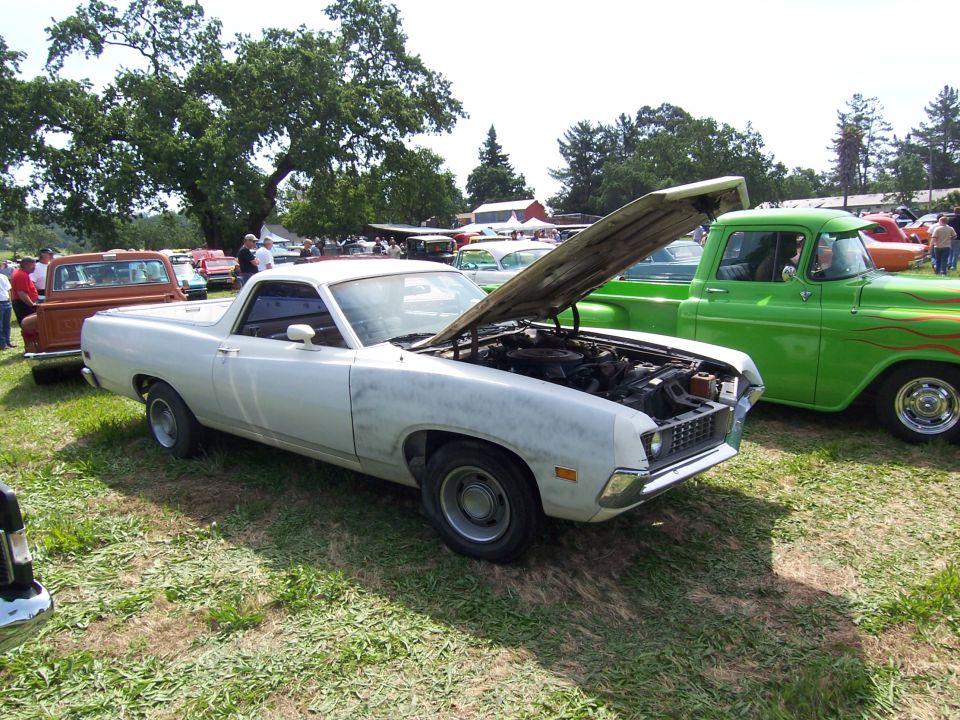 White Ford Ranchero with hood open at a grassy car show, next to a green flamed classic pickup