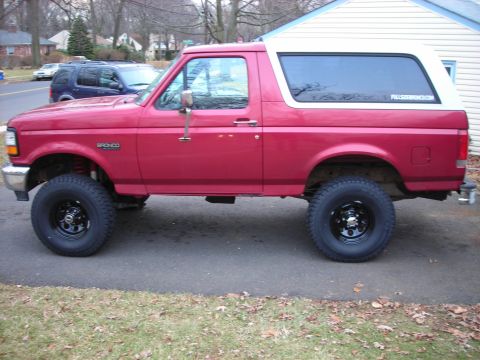 1995 Ford Bronco - A red Ford Bronco with a white removable top is parked on a driveway, showing its lifted suspension and off-road tires.