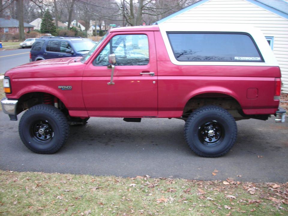 side view of red 1995 Ford Bronco with white top, lifted suspension, and black wheels parked on asphalt