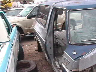1979 Ford Bronco - A blue and silver SUV is seen parked in a junkyard with other vehicles around it.