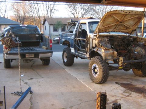 1986 Ford Bronco - A heavily modified truck with its hood up and engine exposed sits next to a pickup truck with a disassembled engine being lowered into its…