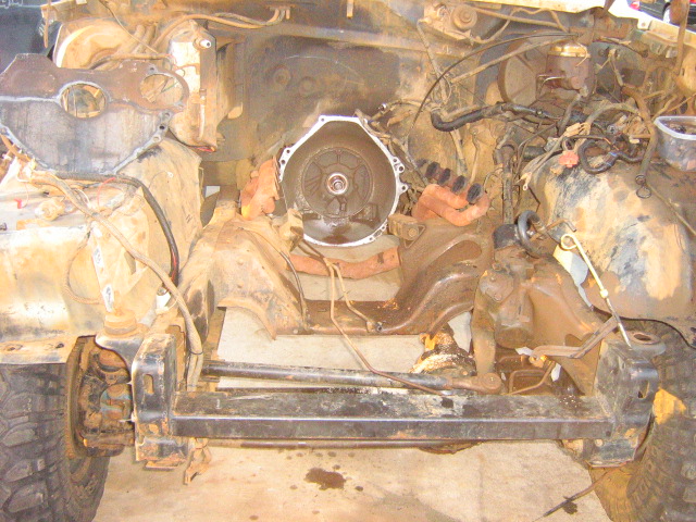 Engine bay of a heavily rusted vehicle, transmission bell housing visible, front tire and suspension components on the left