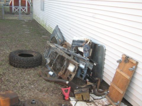 1986 Ford Bronco - A collection of vehicle parts, including a fender, grille, and exhaust system, are piled next to a building.