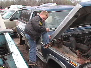 1979 Ford Bronco - A young boy inspects the engine compartment of a blue Ford Bronco in a junkyard setting.