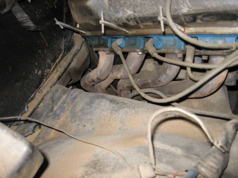 1978 Ford Bronco - Tight, dusty view down the side of a V8 engine in the bay, showing rusty cast iron shorty exhaust headers/manifold runners below a blue-pai…