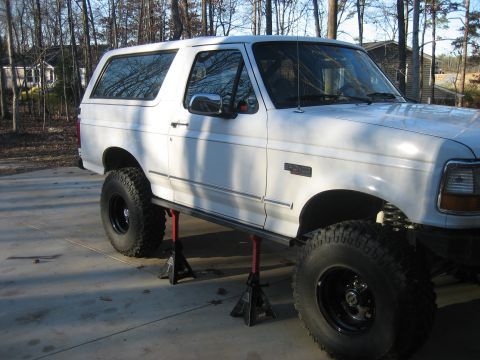 1996 Ford Bronco - Passenger-side profile of a white full-size Ford Bronco raised on red jack stands under its rock slider/nerf bar on a concrete driveway, wi…