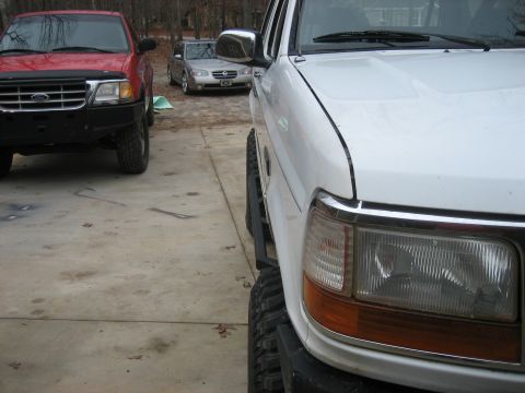 1996 Ford Bronco - Tight close-up of the passenger-side front corner of a lifted white 1990s Ford truck on a driveway, showing the rectangular headlight with…