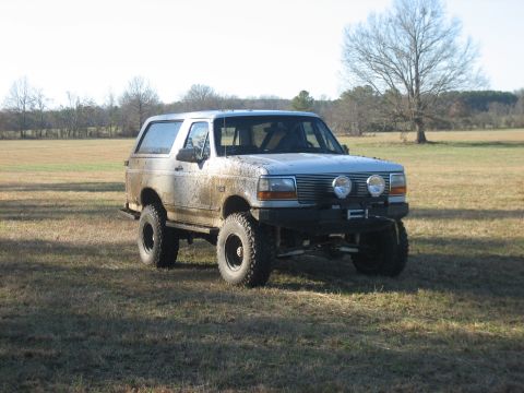 1996 Ford Bronco - Front three-quarter view of a lifted white two-door Ford Bronco splattered with dried mud along the doors and quarter panel, sitting in an…