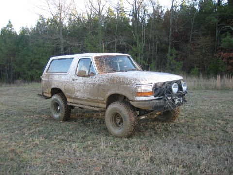 1996 Ford Bronco - White lifted two-door Ford Bronco caked in dried tan mud from rocker to roof, parked in a winter field of dead grass with bare trees behind…