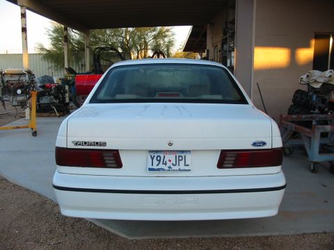 1993 Ford Taurus - The rear of a white 1993 Ford Taurus SHO is visible, parked under a covered area with various engines and equipment in the background.