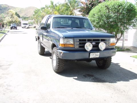 1995 Ford F-150 - A blue SUV with off-road tires and auxiliary lights is parked on a residential street with trees and houses in the background.