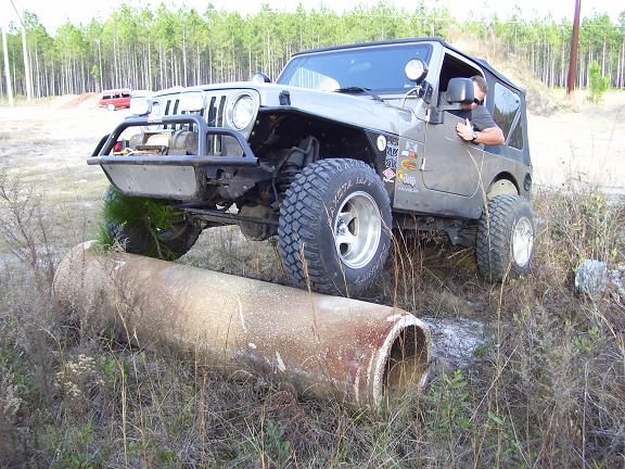 Gray Jeep TJ driving over a large pipe on a dirt path. Driver is visible
