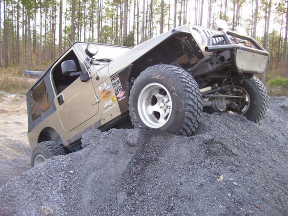 Tan Jeep Wrangler climbing a pile of gravel, front-right wheel elevated