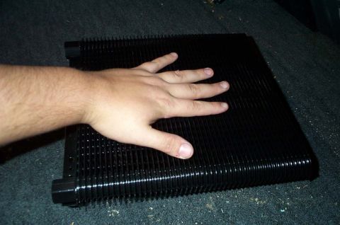1996 Ford Bronco - A hand is placed on a black radiator or heat exchanger, highlighting its ribbed surface and fittings.