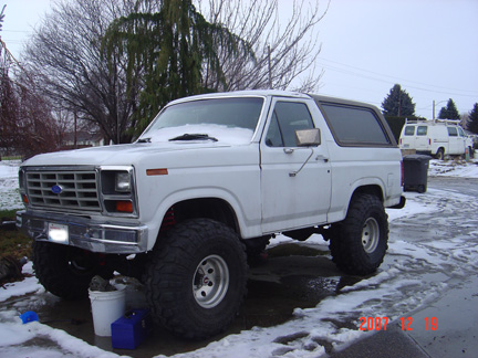 1983 Ford Bronco - A white Ford Bronco with oversized tires sits in a snowy driveway, with two vans visible in the background.