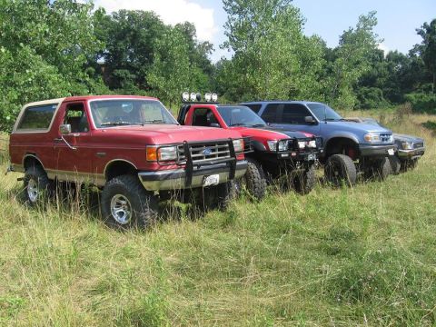 1988 Ford Bronco - A line of modified SUVs and trucks are parked in a grassy area, with the lead vehicle being a red Ford Bronco.