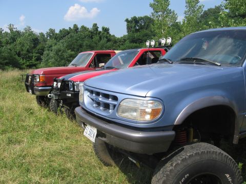 1988 Ford Bronco - A line of SUVs and trucks parked in a grassy field with trees in the background.