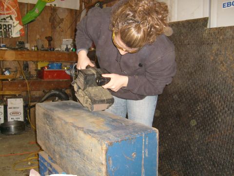 1974 Ford Bronco - A person works on a wooden block with a belt sander in a cluttered garage setting.