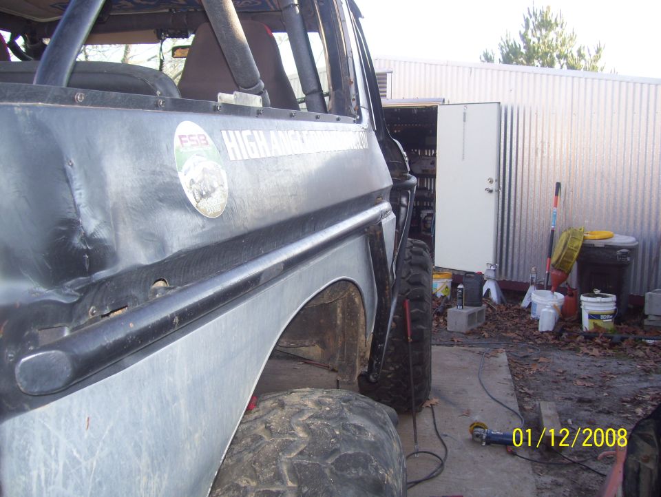 Side view of a modified off-road vehicle with a roll cage and large tires, parked outside a corrugated metal building