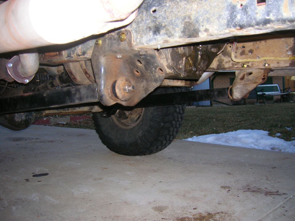 Underbody view of rusty frame, leaf spring, and exhaust pipe. Tire and suspension parts visible