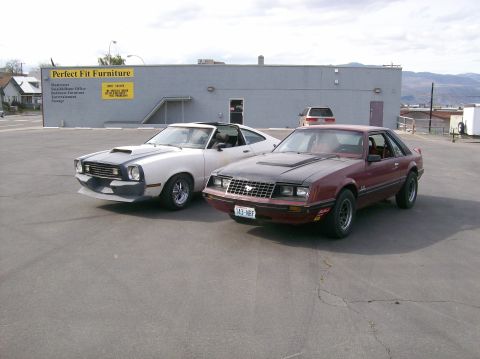 1978 Ford Mustang II - Two classic American coupes, a maroon Ford Mustang II and a white Ford Maverick, are parked in a lot in front of a commercial building.