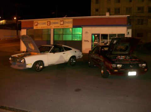 1978 Ford Mustang II - Two cars are parked outside a building with their hoods open, revealing their engines.