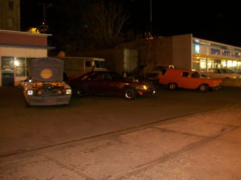 1978 Ford Mustang II - Several vehicles are parked outside a building at night, with two of them having their hoods open.