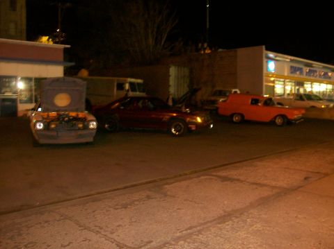 1978 Ford Mustang II - Several vehicles are parked in a lot at night, with one car's hood open.