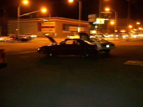 1978 Ford Mustang II - Two cars are parked on a street at night, one with its hood up.