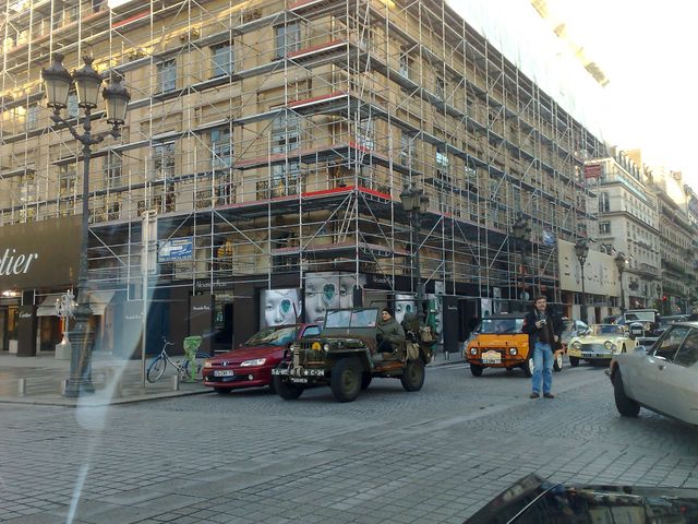 Several cars and a military-style jeep parked on a cobblestone street in front of a building covered in scaffolding