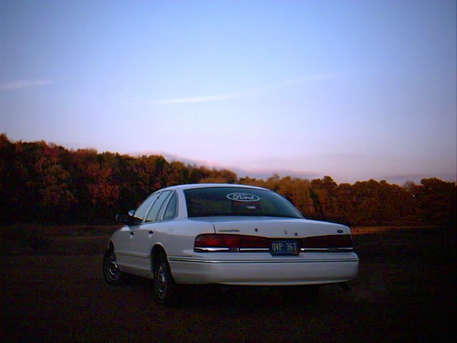 White Ford Crown Victoria sedan, rear 3/4 view, parked in a field under a pink dusk sky