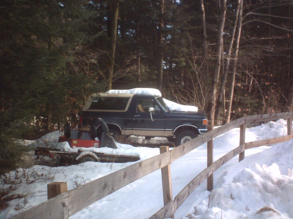 Dark blue Ford Bronco with tan trim parked in snow next to a trailer with a snowblower