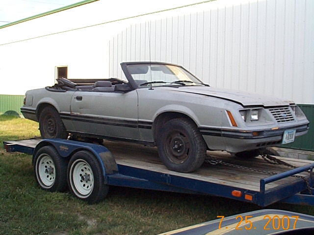 Silver Ford Mustang convertible project car on a blue flatbed trailer beside a white metal building